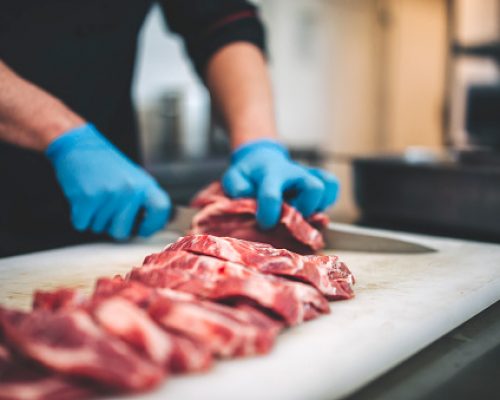 closeup view of a young caucasian man cutting raw pork neck with sharp knife. He makes steaks for grill in restaurant kitchen